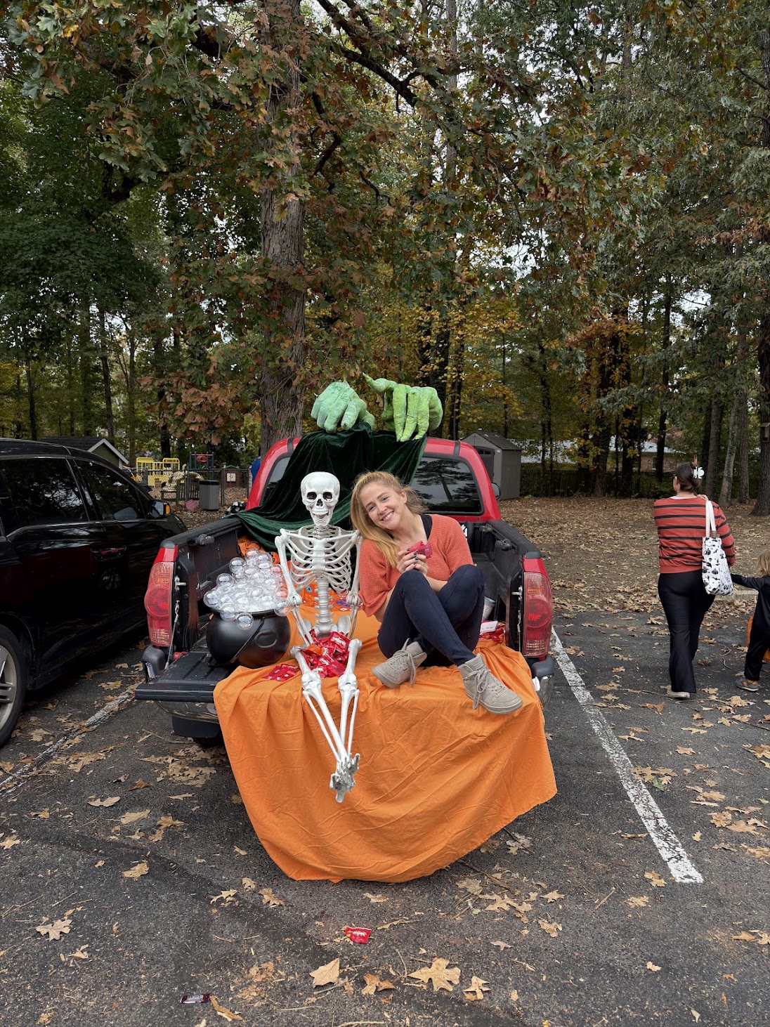 A Welborne volunteer smiling from the tailgate of a red pickup truck decorated for Trunk or Treat with a full-length skeleton, a cauldron of candy, and giant green monster hands